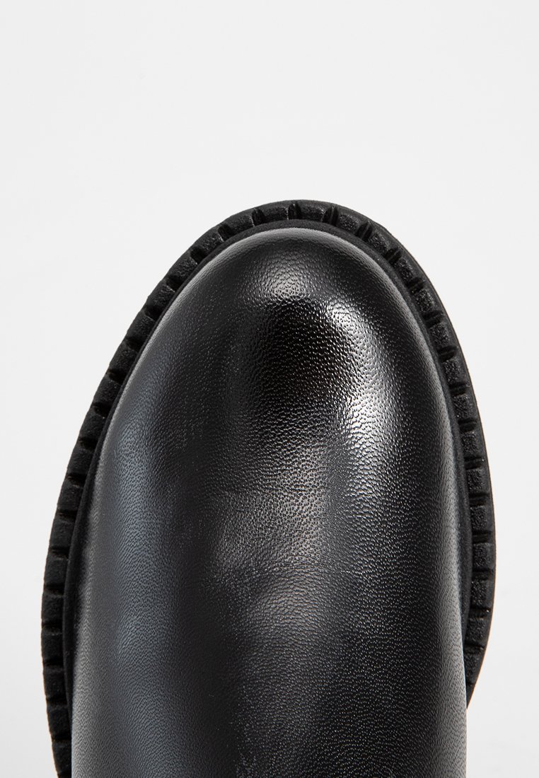 Bota de cuero negro con superficie suave y punta redondeada. Suela texturizada con un detalle de borde cosido. Sin herrajes ni acentos visibles.