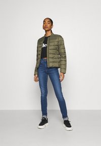 Casaco puffer verde-azeitona com fecho éclair, textura matelassada, punhos canelados, usado com um top preto cropped e jeans skinny azuis.