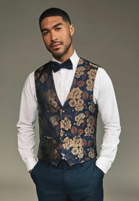 Marineblaues, floral gemustertes Weste mit goldenen und braunen Akzenten, getragen über einem weißen Hemd, kombiniert mit einer schwarzen Fliege und zwei vorderen Taschen.