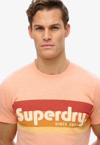 Lyserød bomulds t-shirt med et iøjnefaldende grafisk print i rød, orange og gul, der læser "Superdry" og "siden 2003" på tværs af brystet.
