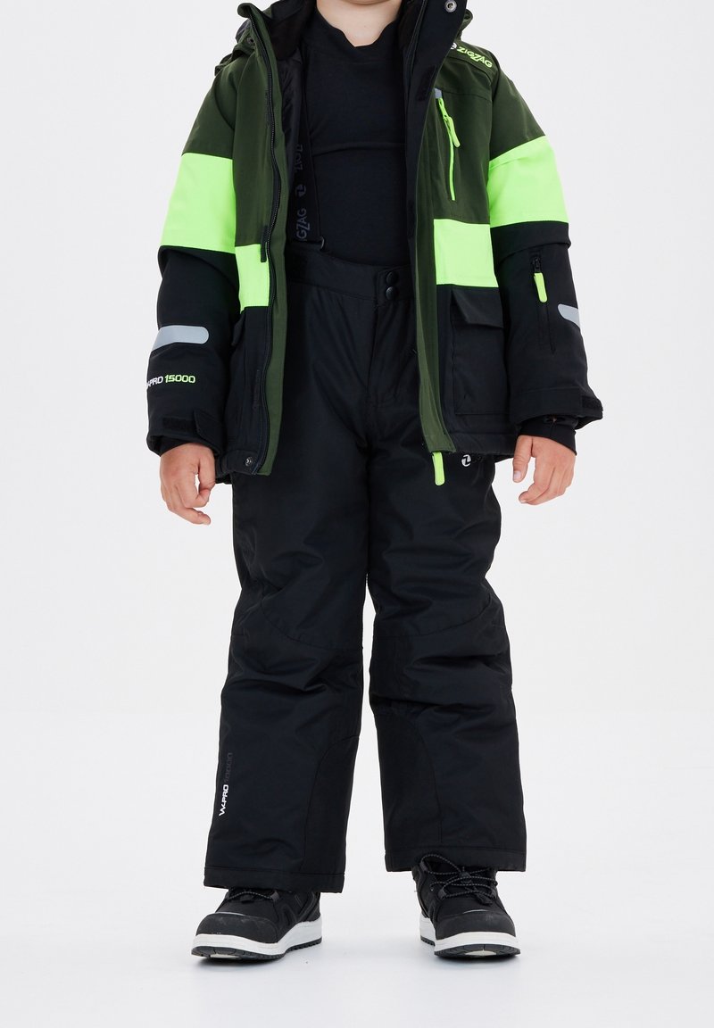 Enfant portant un pantalon de ski noir et une veste d’hiver verte et noire avec des accents jaune néon, debout, les mains détendues le long du corps.