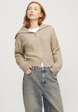 Beige geribbelde brei-cardigan met een grote capuchon, ritsluiting en lange mouwen, gecombineerd met high-waisted vervaagde blauwe jeans.