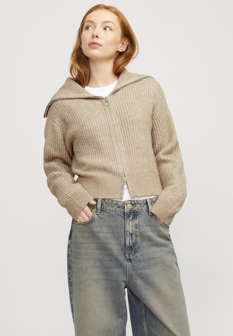 Beige ribbad stickad kofta med en stor huva, dragkedja och långa ärmar, kombinerad med högmidjade blekta blå jeans.