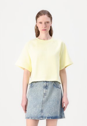 Felpa gialla cropped con maniche corte, abbinata a una gonna di jeans con branding bianco ripetuto. Design semplice con una vestibilità rilassata.