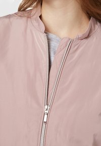 Lys pink bomberjakke med høj krave, sølv lynlås og glat, let skinnende stof. Har et simpelt, rent design uden mønstre.