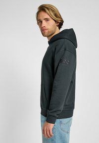 Sudadera gris oscuro hecha de una tela suave, con un bolsillo tipo canguro, puños acanalados y un logo bordado en la manga. Se lleva con jeans azul claro.