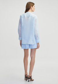 Helles blaues Langarmhemd aus durchscheinendem Stoff, mit einer Rückenpasse und Knöpfen an den Ärmelbündchen, kombiniert mit passenden blauen Shorts.