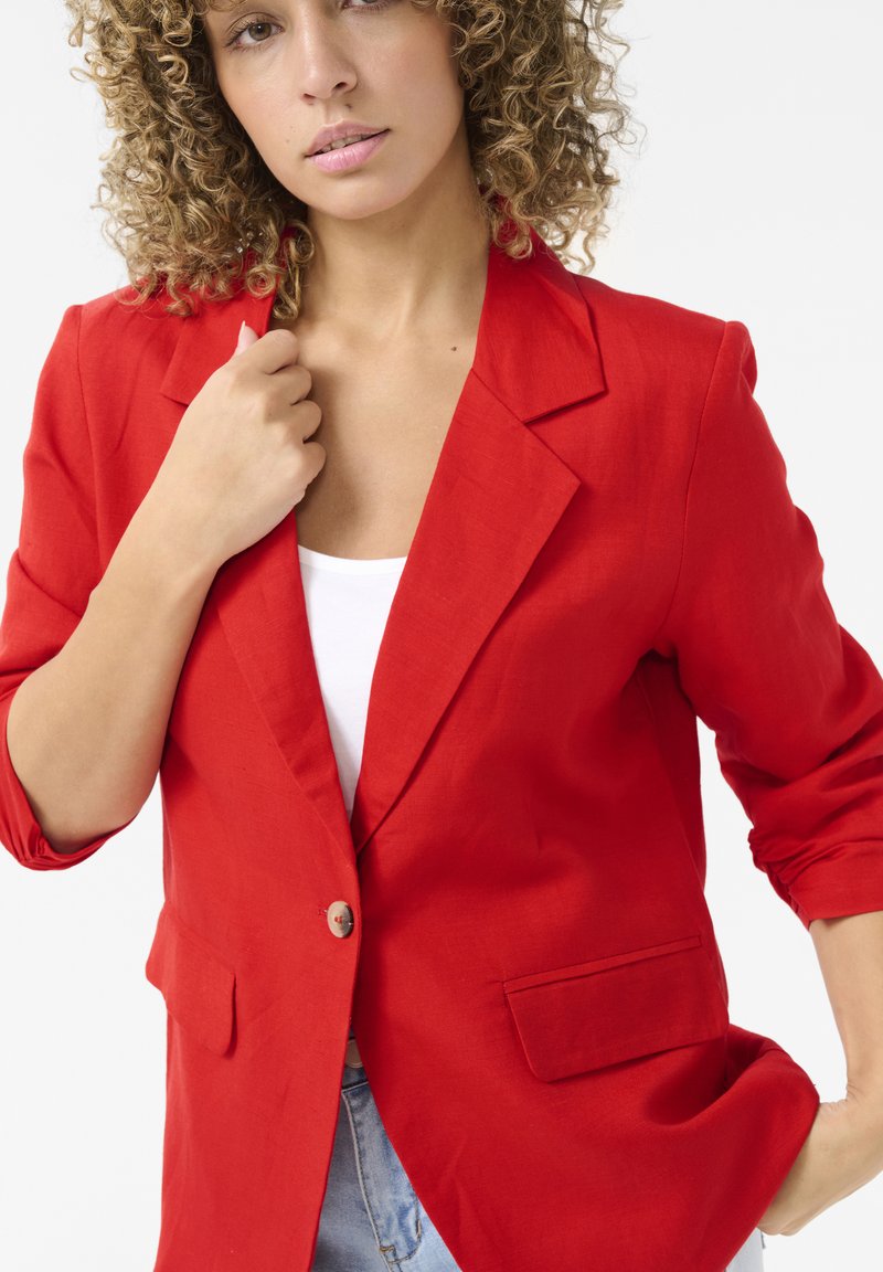 Blazer en lin rouge avec un col à revers cranté, une fermeture à un bouton et deux poches. Manches retroussées et texture lisse. Associé à un haut blanc.