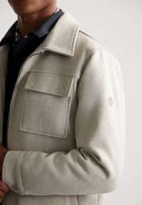 Gray fleece jacket with a collar, flap pocket on the chest, and subtle texture. Features a logo detail on the sleeve and a zipper closure.