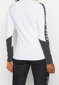 Witte sportieve top met lange mouwen en grijze accenten, met een gebogen naadd ontwerp en het "PEAK PERFORMANCE" logo langs de mouw.