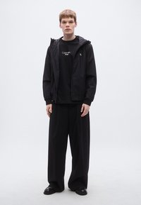 Μαύρο ελαφρύ μπουφάν με κουκούλα, φορεμένο πάνω από μαύρο μπλουζάκι με λευκό κείμενο λογοτύπου, συνδυασμένο με φαρδιά μαύρη παντελόνα και μαύρα παπούτσια.