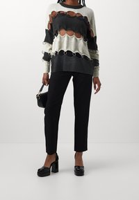 Pull tricoté noir et blanc avec découpes circulaires et anneaux métalliques, porté avec un pantalon noir et des chaussures à plateforme. Sac à main en main.