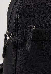 Gros plan sur la poche avant d'un sac à dos en cuir noir texturé avec une fermeture éclair et une languette de tirage sur un fond uni.