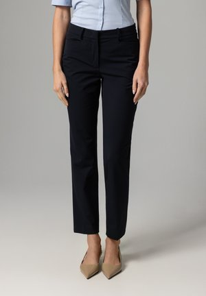 Frau trägt eng anliegende schwarze Stoffhose, hellblaues Hemd mit Knöpfen und beige spitze Pumps, steht vor einem schlichten grauen Hintergrund.