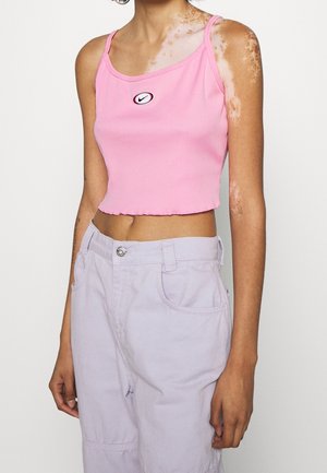 Person med vitiligo iført en lyserød cropped tanktop med et Nike-logo og lyse lavendel-højtaljede bukser mod en ensfarvet baggrund.