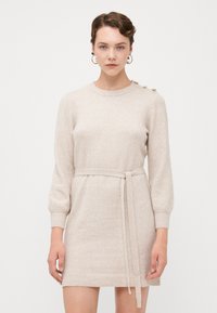 Strikket kjole i lys beige, med lange ermer, en ribbestrikket kant, et belte i livet og dekorative knapper på den ene skulderen.