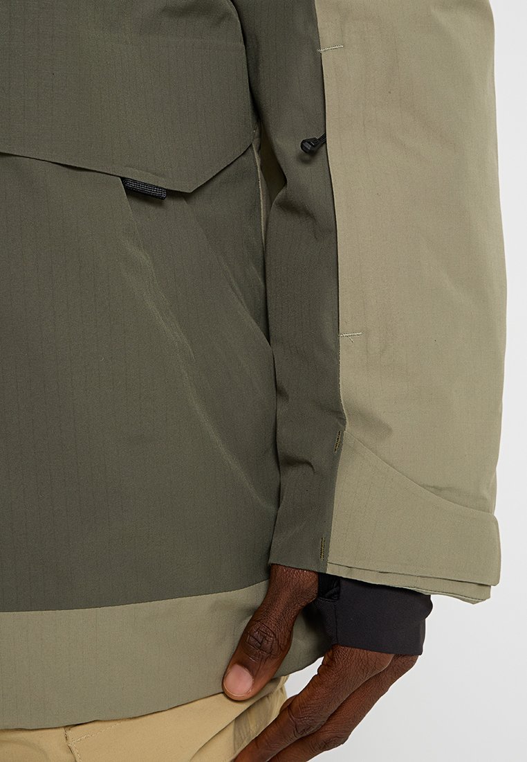 Una chaqueta bicolor con una sección superior de color verde oscuro y una sección inferior de color verde claro. Tela texturizada con costuras visibles y puños ajustables.