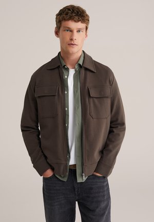 Bruine jas met ritssluiting en twee voorzakken, gecombineerd met een groen overhemd met knopen over een wit T-shirt. Donkere spijkerbroek maakt de outfit compleet.