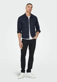 Giacca di jeans blu navy con due tasche sul petto, colletto classico e bottoni metallici, abbinata a pantaloni neri e sneakers bianche. Design semplice e moderno.