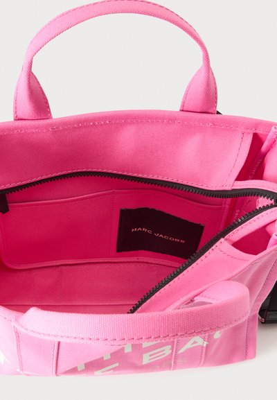 Sac cabas en tissu rose vif avec des poignées courtes, fermeture éclair ouverte dévoilant une poche intérieure et une étiquette noire avec le texte « Marc Jacobs ».