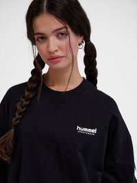 Mladá žena s dlouhými zapletenými vlasy, která má na sobě stříbrné kruhové náušnice a černou mikinu značky Hummel na bílém pozadí.