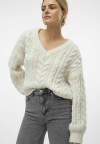 Un pull en maille torsadée épaisse avec un décolleté en V et de longues manches côtelées, associé à un jean taille haute gris foncé. Texture douce et coupe décontractée.