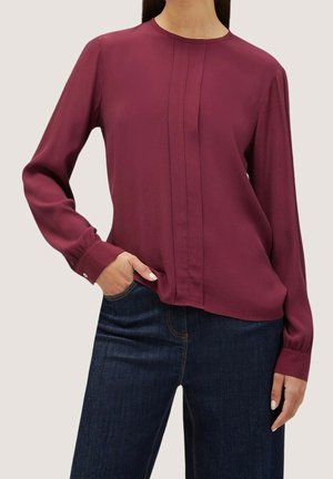 Bordeauxrode blouse met lange mouwen, voorzien van een ronde hals, plooitjes aan de voorkant en manchetten met knopen, gecombineerd met een donkere spijkerbroek.