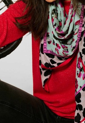 Femme portant un pull rouge et une écharpe bleu clair avec des motifs floraux roses et noirs, assise sur une chaise avec un accoudoir en spirale.