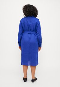 Abito camicia in lino blu con vita cintata, maniche lunghe e lunghezza fino al polpaccio. La vista posteriore mostra un design e una texture semplici e puliti. Calzature nere.