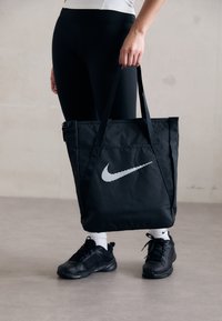 Persoon in zwarte leggings, zwarte sneakers en witte sokken, met een zwarte Nike draagtas met een witte swoosh logo.