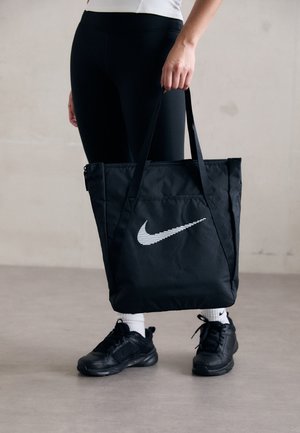Persoon in zwarte leggings, zwarte sneakers en witte sokken, met een zwarte Nike draagtas met een witte swoosh logo.