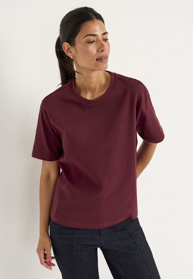 Vrouw met donker haar in een paardenstaart, in een effen bordeauxrode T-shirt met korte mouwen en donkere jeans, staand tegen een effen witte achtergrond.