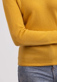 Gul stickad tröja med långa ärmar, åtsittande design och ribbade muddar, i kombination med ljusblå denimjeans. Mjuk textur synlig.