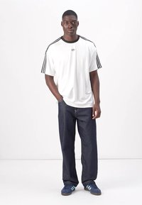 Witte t-shirt met korte mouwen en zwarte kraag, voorzien van drie zwarte strepen, gecombineerd met donkerblauwe baggy jeans en marineblauwe Adidas sneakers.