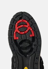 Zwarte rubberen schoenzool met een gestructureerd patroon, met rode ronde hardware in het midden en gele merkopdruk nabij de hiel.