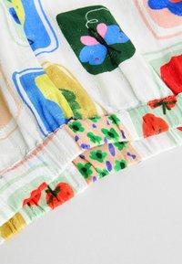 Färggörande mönstrad tyg med blommor och fjärilar, med en rynkad nederkant. Materialet verkar lätt och texturerat, på en vit bakgrund.