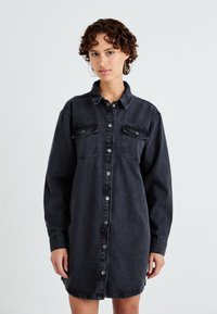 Zwarte denim jurk met lange mouwen, knoopsluiting aan de voorkant, twee borstzakken en een relaxte pasvorm, met een gladde textuur en lichte slijtage.