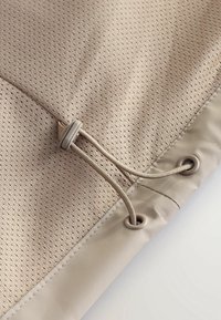 Tissu beige avec de petites perforations, doté d'un cordon beige passé à travers deux œillets métalliques avec un bouton-pression en plastique réglable.