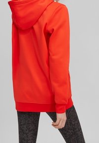 Personne portant un sweat à capuche rouge vif et des leggings sombres à motifs sur un fond gris clair uni.