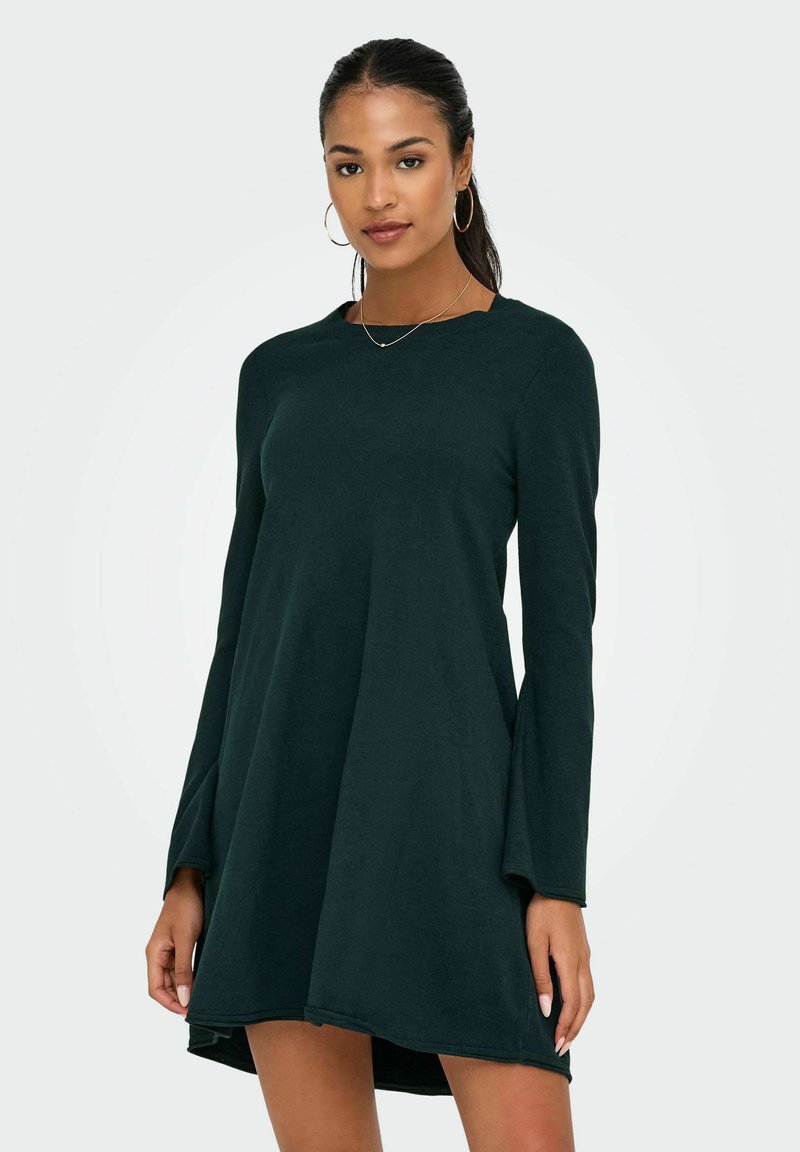 Donna che indossa un abito lungo verde scuro a linea ad A con maniche a campana, orecchini a cerchio e una delicata collana, in piedi davanti a uno sfondo neutro.