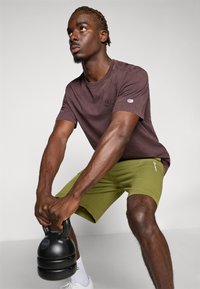 Braunem Baumwoll-T-Shirt mit kleinem Logo, kombiniert mit grünen Shorts. Die Person hält eine schwarze Kettlebell, mit weißen Turnschuhen sichtbar.