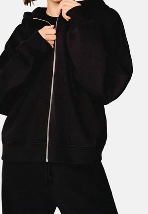 Personne portant un sweat à capuche noir surdimensionné zippé et un pantalon noir, mains près de la fermeture éclair au niveau de la poitrine, fond neutre.