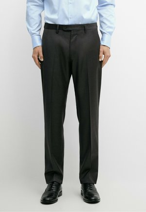 Homme portant un pantalon de costume noir, des chaussures habillées noires et une chemise à manches longues bleu clair, debout devant un fond uni.