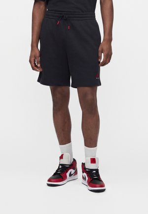 Mann trägt schwarze Jordan-Shorts mit roten und schwarzen Air Jordan Sneakers und weißen Socken, steht vor einem einfachen weißen Hintergrund.