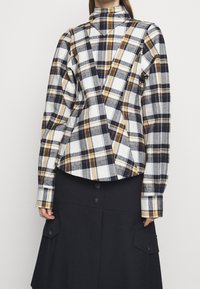 Plaid shirt in white, navy, and gold with a high collar and unique asymmetric cut. Features buttoned cuffs and a textured fabric.