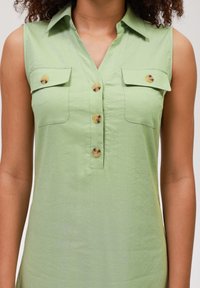 Canottiera verde senza maniche con bottoni frontali, due tasche sul petto e grandi bottoni rotondi. Il materiale sembra leggero e texturizzato.
