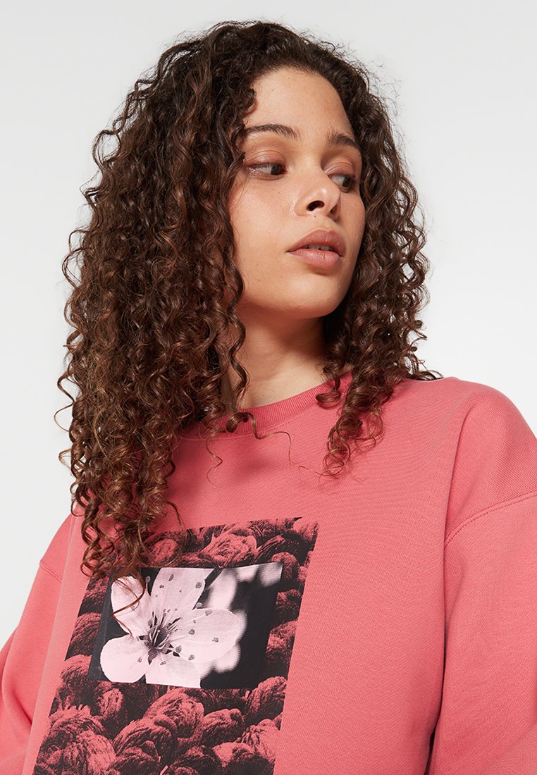 Różowy sweter z miękkiego materiału, z dużą graficzną florystyczną aplikacją z przodu. Luźny krój z prążkowanym dekoltem i mankietami.