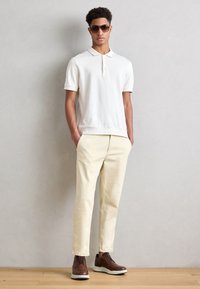 White polo shirt with short sleeves and a collar, paired with cream-colored trousers and brown lace-up shoes with a white sole. Dark sunglasses.