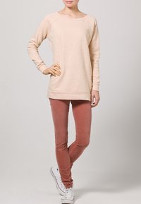 Broskvově zbarvený top s dlouhým rukávem, volného střihu a kulatým výstřihem, doplněný úzkými rezavými kalhotami a bílými teniskami.