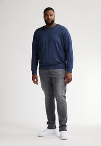 Marineblauwe langemouwen trui in een textuurgebreide stof, gecombineerd met grijze slim-fit jeans en witte sneakers, staand tegen een neutrale achtergrond.